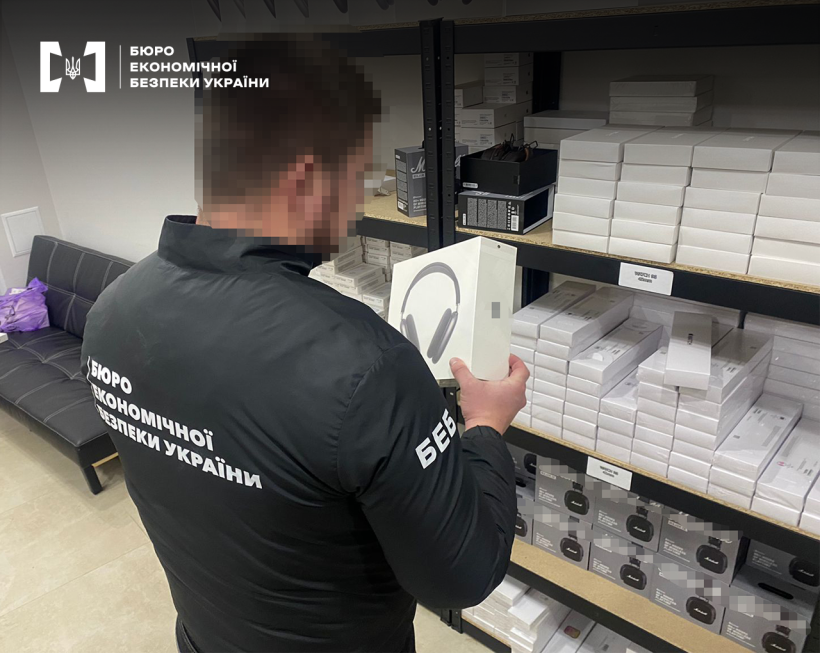 Слідчі дії БЕБ. Детектив оглядає коробку з навушниками. На полицях велика кількість товарів у пакованні.