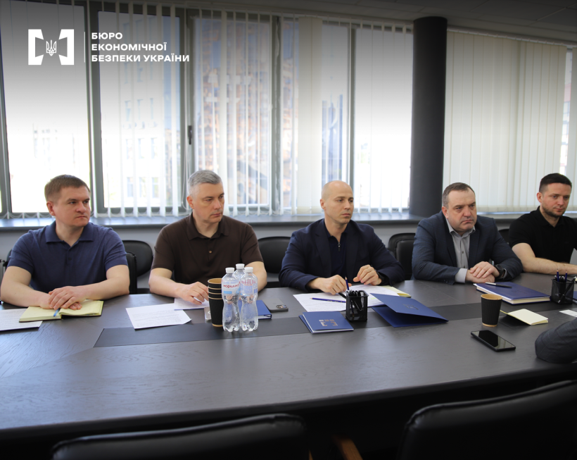 Учасники зустрічі за столом. На столі документи, записники, папки і блокноти з логотипом БЕБ, олівці й ручки у підставках, ПЕТ пляшки з водою, стакани.