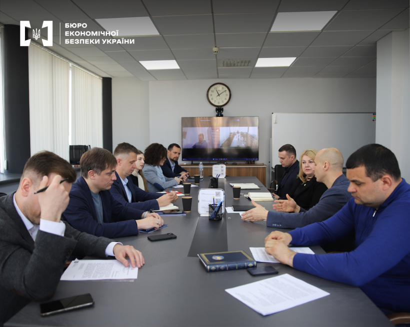 Учасники круглого столу БЕБ. Сучасний зал, на столі документи, записники, смартфони. Подалі настінний годинник, екран, фліпчарт.