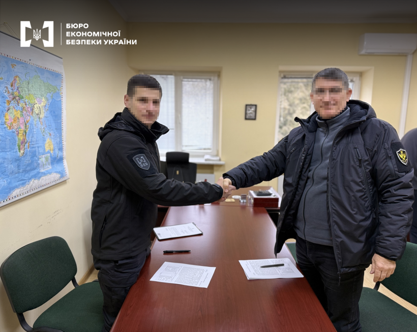 Рукостискання працівників БЕБ та АРМА, обличчя заблюрено. Офісне приміщення, стіл для нарад, стільці, на стіні карта світу.