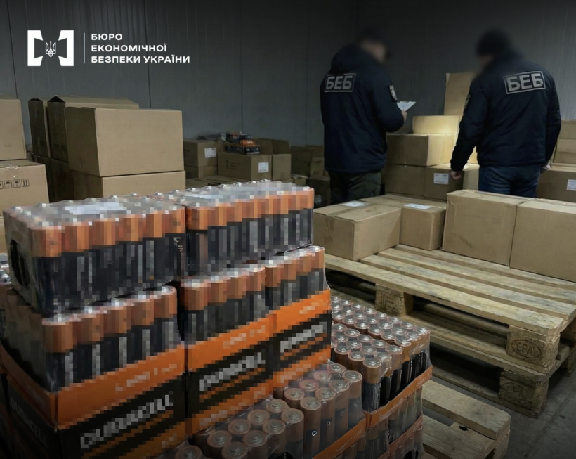 Детективи БЕБ проводять обшук в складському приміщенні заповненому стосами коробок. На передньому плані прозорі упаковки з лужними батарейками