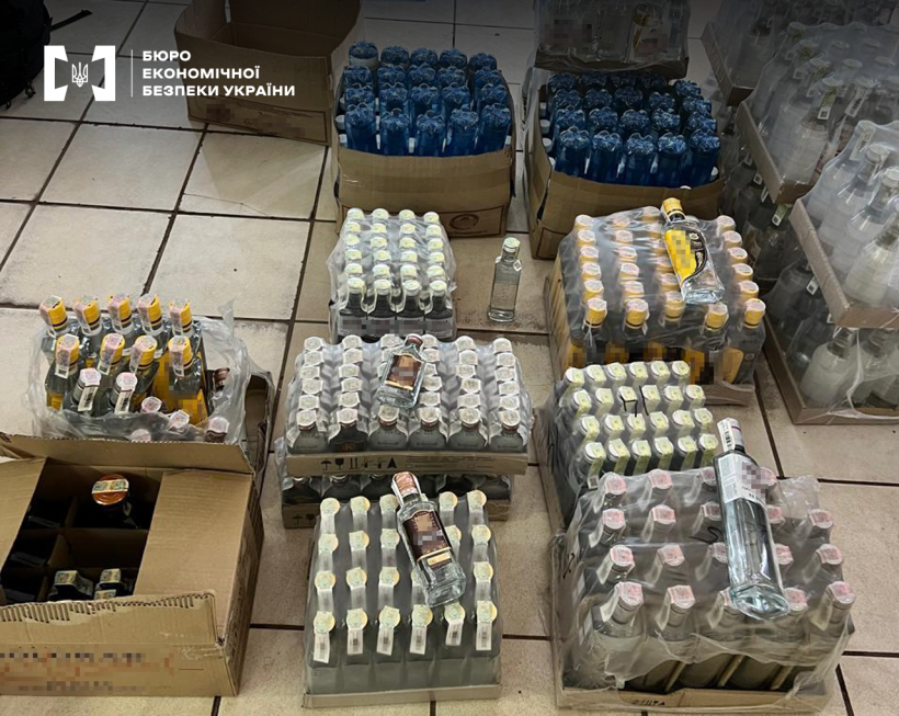 Контрафактний алкоголь у прозорих скляних та синіх ПЕТ пляшках запакований плівкою та у картонних ящиках на підлозі.