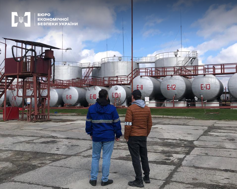Детективи БЕБ на території нафтопереробного заводу оглядають резервуари з написами від Е-6 до Е-13. Далі нафтові баки на фоні блакитного неба з хмарами.