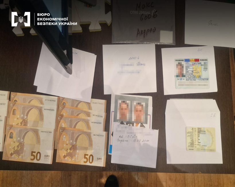 На поверхні столу розкладені купюри номіналом 50 євро, фото на документи, нотатки на білих папірцях, конверти, пластикові ID картки.