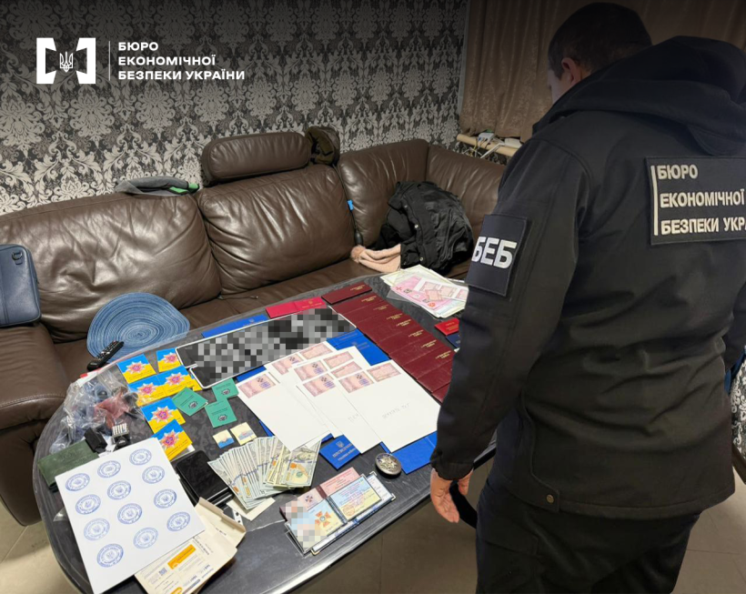 Детектив БЕБ під час обшуку проводить огляд речових доказів - документи, посвідчення, бланки, печатки, валюта, що розкладені на столі біля дивану.