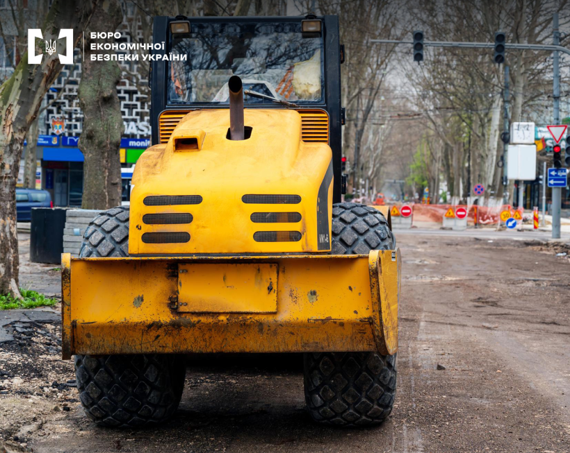 Ремонтні роботи на дорозі: жовтий дорожній каток великим планом, далі перекрита дорога, світлофори, багато дорожніх знаків, дерева.