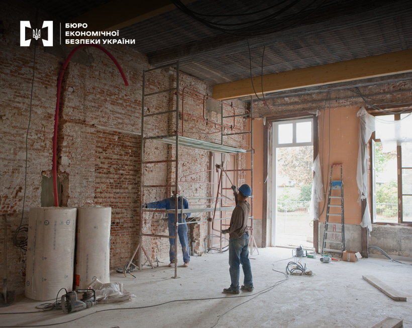 Двоє робітників проводять оздоблювальні роботи всередині приміщення. Біля цегляної стіни стоїть риштування, на підлозі - будівельні матеріали, інструменти, електричний подовжувач
