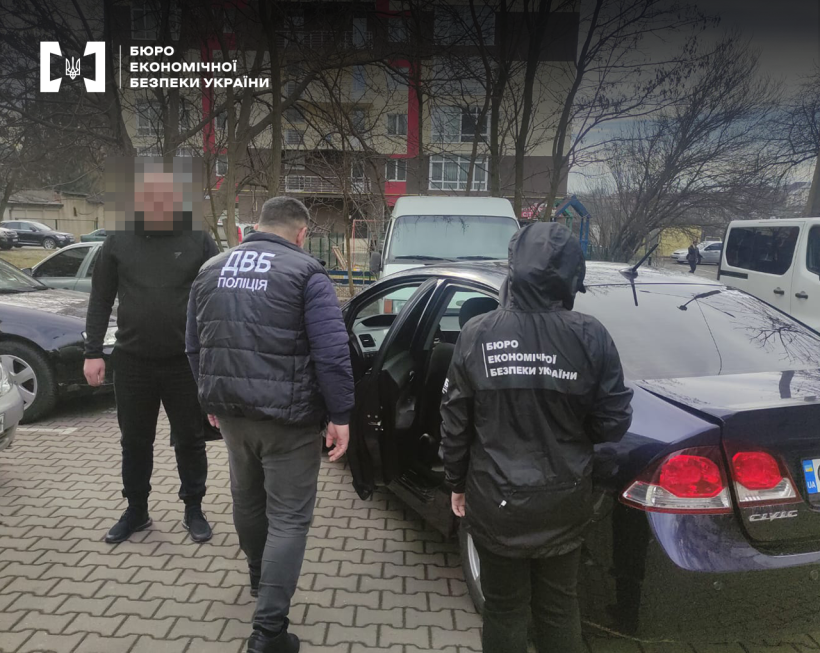 Автостоянка. Детектив БЕБ та працівник ДВБ Поліції проводять обшук автомобіля. Поруч багатоповерхівка, дерева.