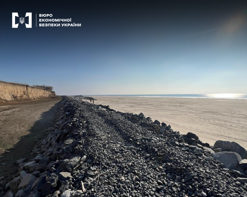 Великий насип будівельного каміння вздовж піщаного берегу. Водосховище та сонячне небо на горизонті.