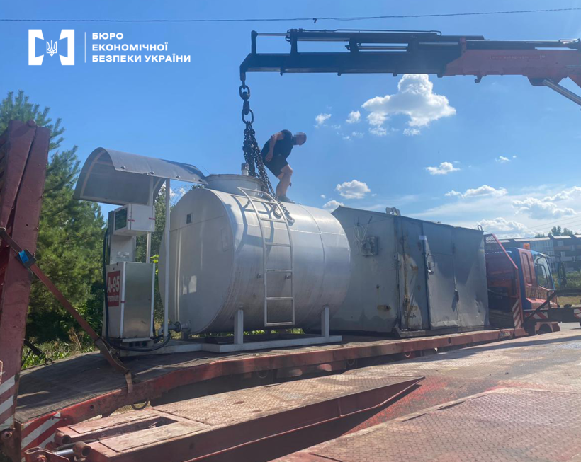 Завантаження підйомним краном пальнороздавальної колонки з резервуаром на причеп вантажівки. На резервуарі для пального чоловік на тлі блакитного неба