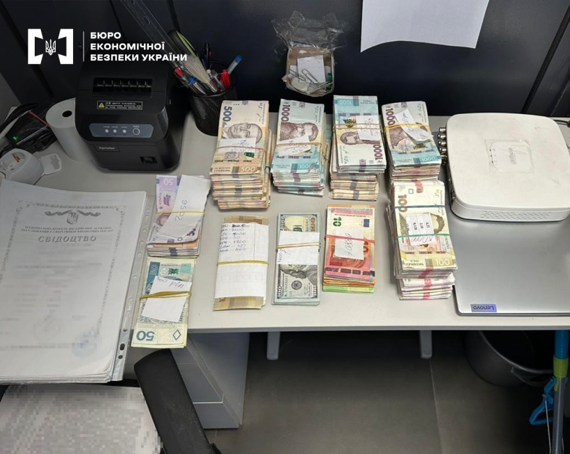 Пачки гривень, доларів США, євро та злотих різних номіналів на офісному столі. Поруч документи,  офісне обладнання, ноутбук та модем.