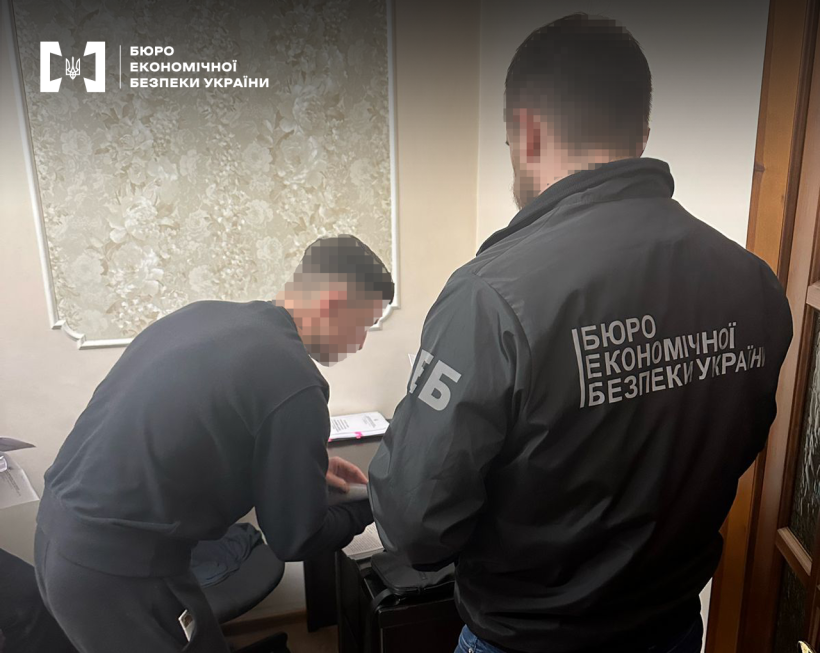 Детектив БЕБ ознайомлює фігуранта справи з підозрою. Обличчя обох чоловіків заблюрено.
