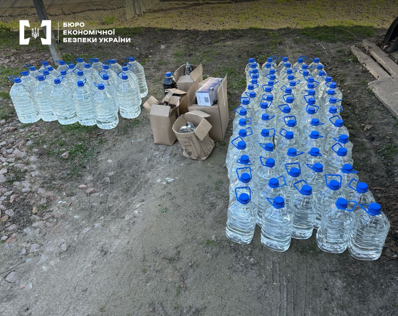 Велика кількість ПЕТ пляшок 5 і 6 літрів з прозорою рідиною, що стоять на землі розсортовані за об’ємом. Між ними кілька розкритих картонних коробок