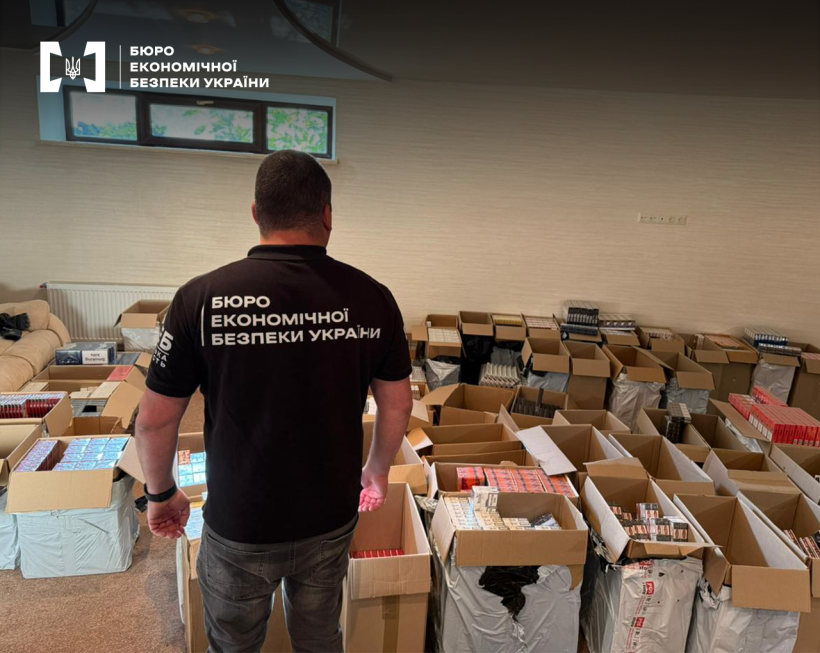 Детектив БЕБ оглядає кімнату з великою кількістю відкритих картонних коробок, що містять блоки сигарет різних марок.