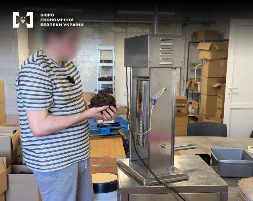 Чоловік із грудкою тютюну в руках стоїть біля фасувального верстата на тлі картонних коробок та стелажів