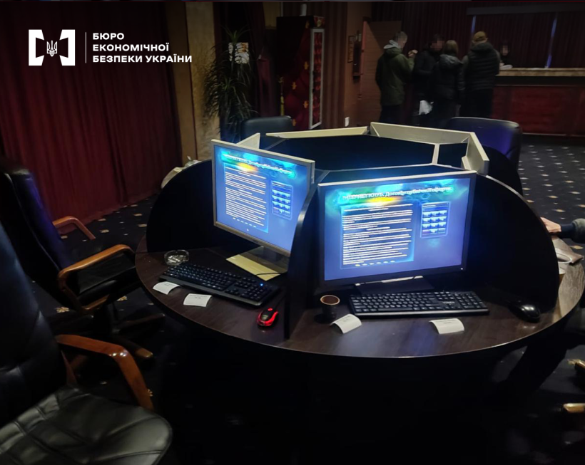 Круглий стіл з увімкненими комп'ютерами у темному приміщенні грального закладу.