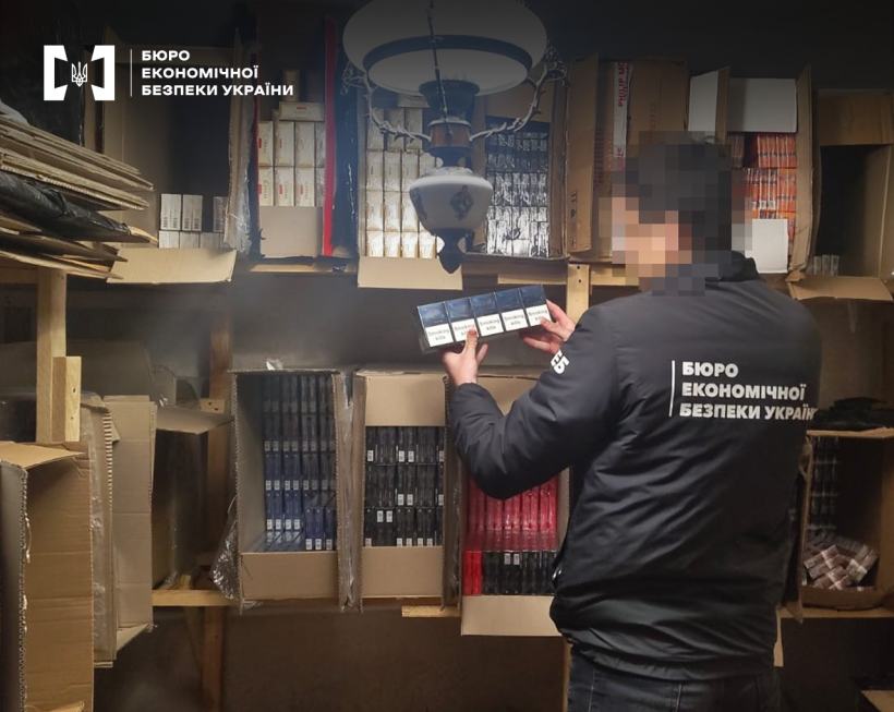 Детектив БЕБ проводить обшук приміщення, де фігурант справи зберігав сигарети. На полицях картонні коробки з сигаретами у блоках різного кольору.