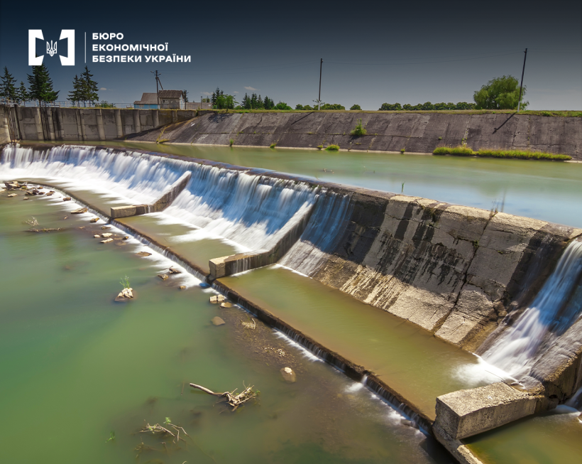 Бетонний водоскид з багатоярусним каскадом, вода стікає сходинками в каламутну річку. На горизонті дерева, хати, блакитне небо