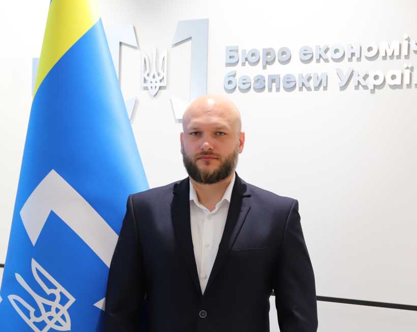 Директор Бюро економічної безпеки України Олександр Цивінський біля прапору з логотипом БЕБ на білому тлі з написом: «Бюро економічної безпеки»