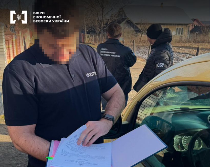 Працівник прокуратури вивчає документи. Обличчя заблюрено.  Поруч автомобіль, за ним працівники БЕБ та СБУ. Далі земельна ділянка, одноповерхові житлові будинки