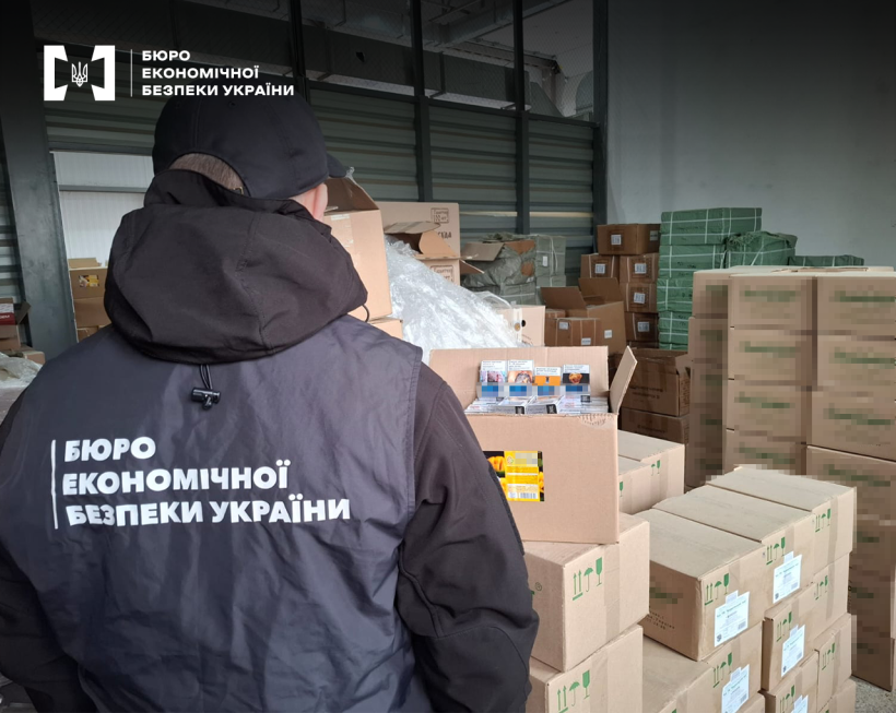 Безліч картонних коробок, що стоять рядами одна на одній. Одна коробка відкрита, в ній сигарети. Детектив БЕБ проводить огляд товару.