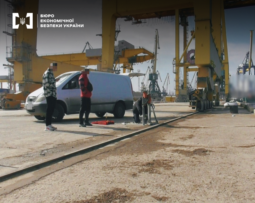 Територія порту з великою кількістю жовтих портових кранів. Біля білого мінівену троє чоловіків, один з яких проводить заміри спеціальним приладом.