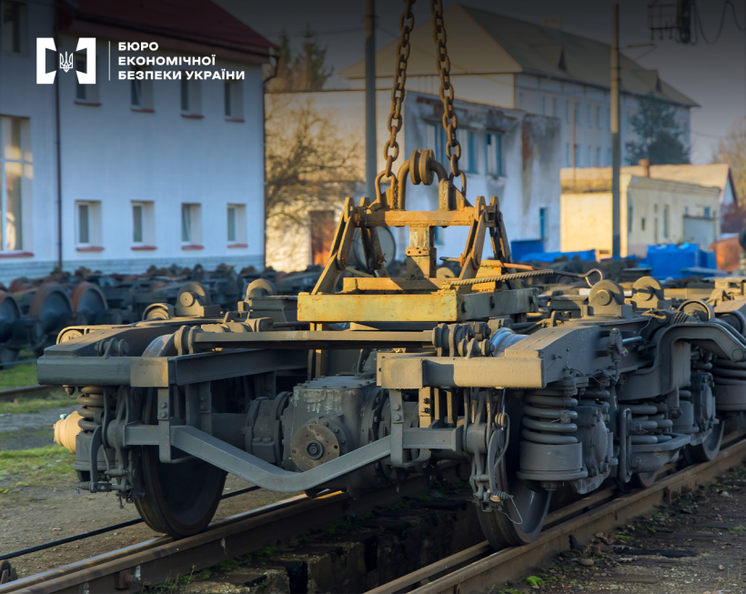 Залізничний візок, під'єднаний до крана, на ремонтній ділянці. На задньому плані — промислові будівлі та колеса під ними.