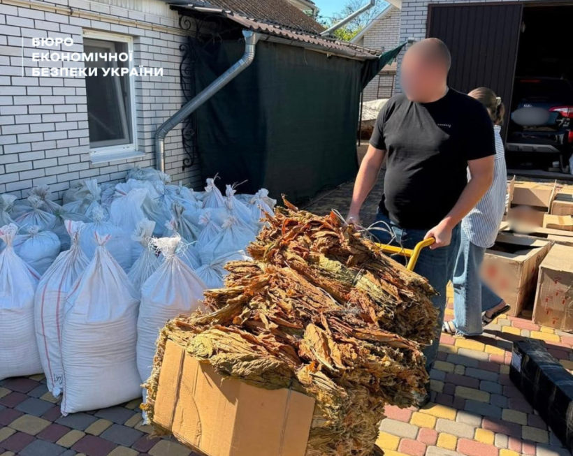 A large pile of yellow-brown tobacco leaves in a box on a wheelbarrow next to white sacks near a brick house.
