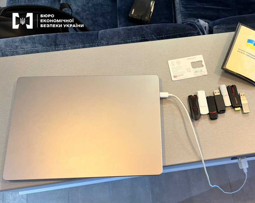 Сірий ноутбук, 7 USB флешок різних дизайнів, упаковка від сім-картки, паспорт громадянина України на  сірому столі.