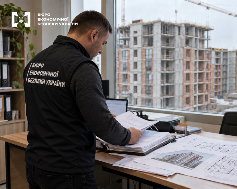 A ESBU detective examines construction blueprints in an office against the background of a high-rise building under construction outside the window.