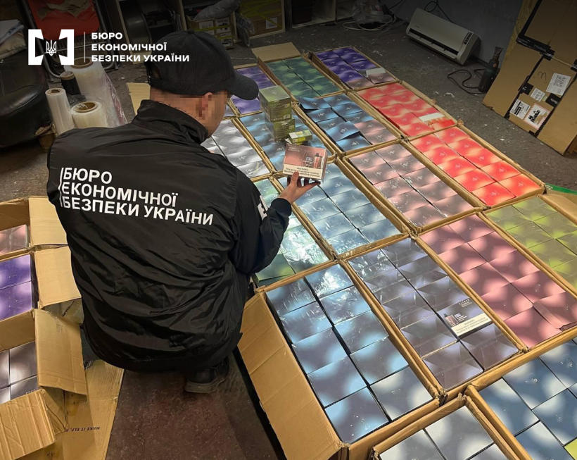 Велика кількість відкритих картонних коробок заповнених одноразовими електронними сигаретами різних смаків. Детектив БЕБ проводить огляд.