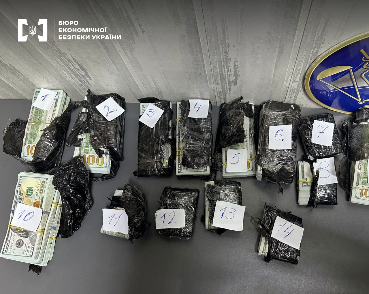 Пачки стодоларових банкнот у розірваних чорних мішках, обмотаних скотчем. На кожній пачці прикріплений папірець із цифрами від 1 до 14.