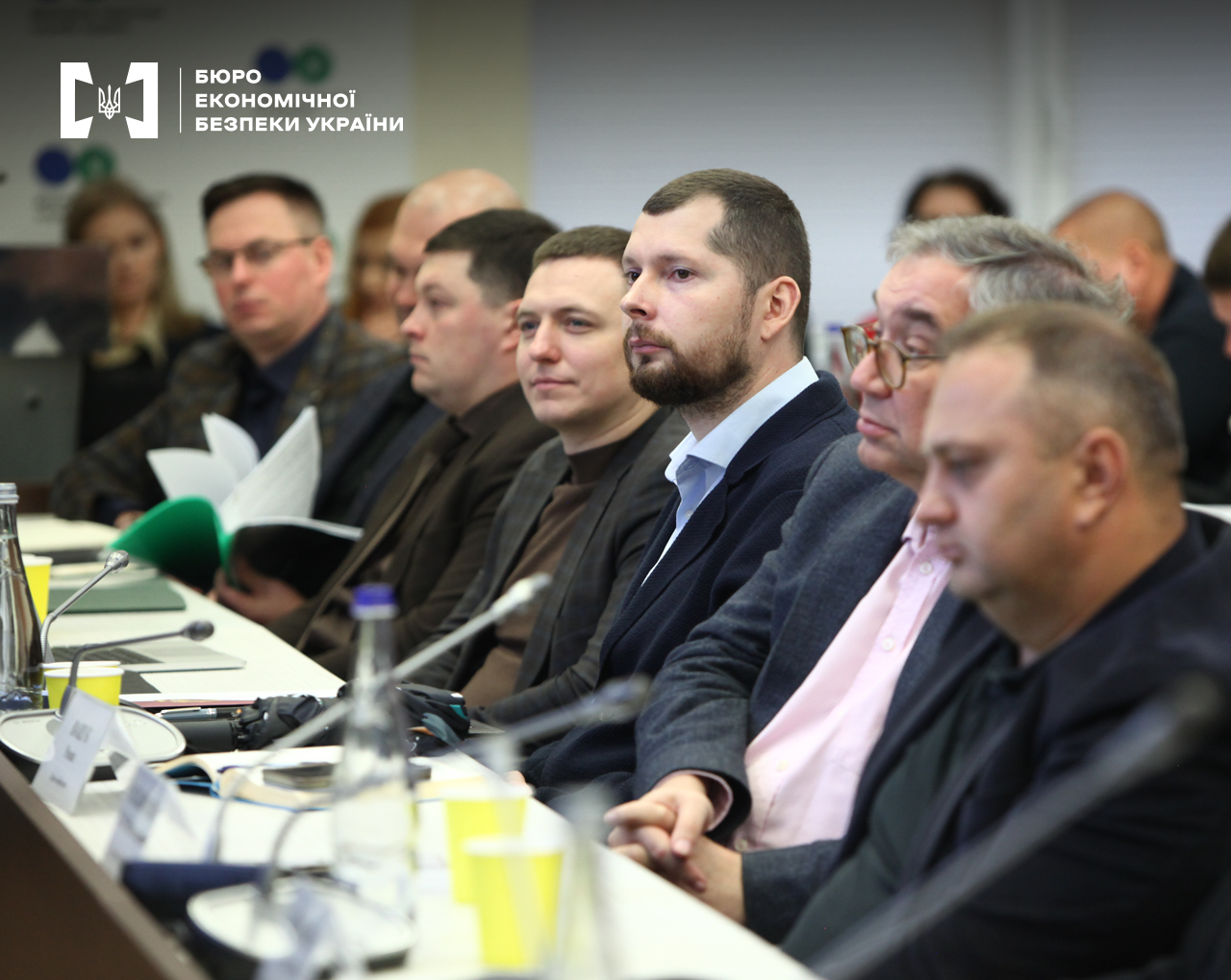 Учасники зустрічі сидять за довгим столом у конференц-залі. На столі мікрофони, вода в скляних пляшках, стакани, нотатники