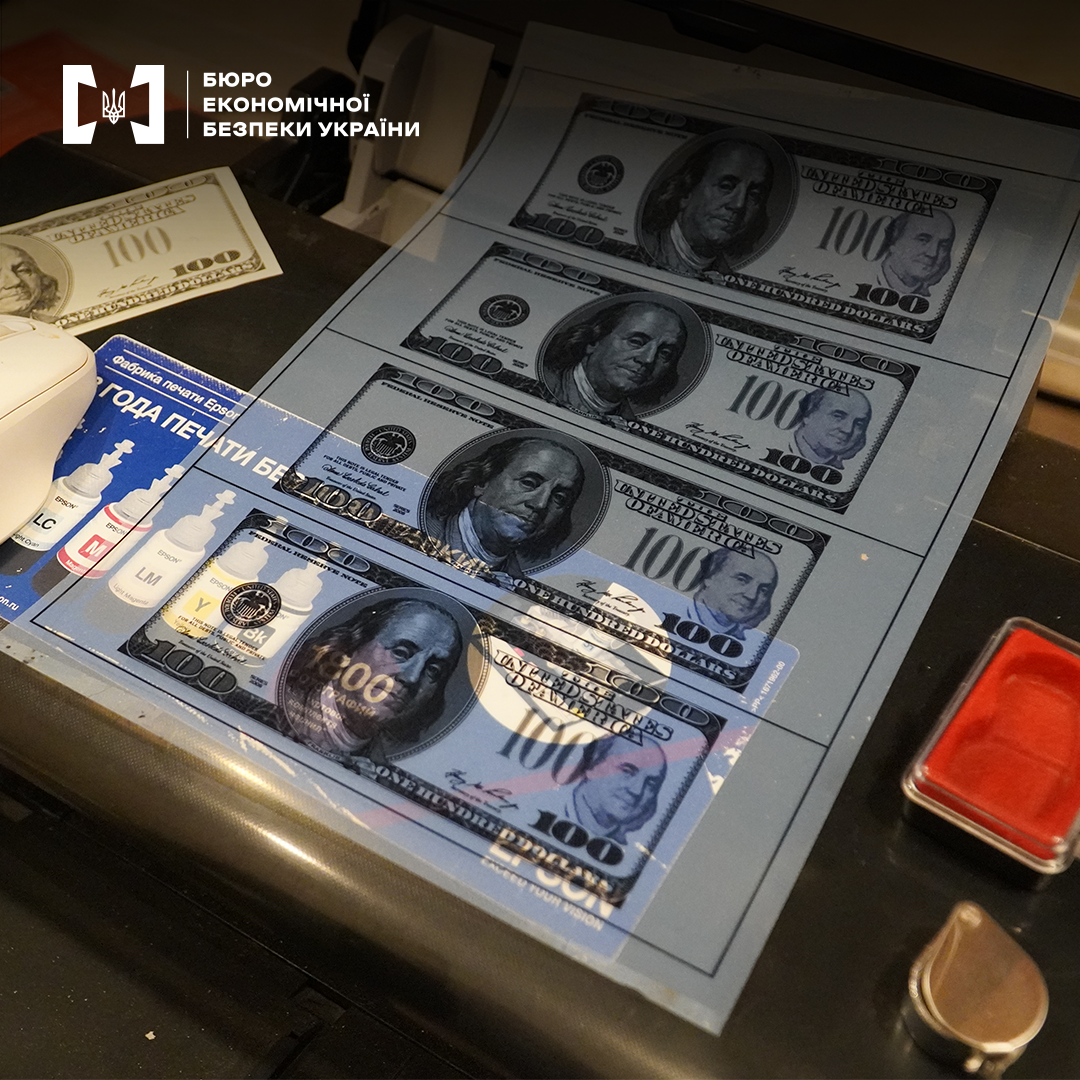 Аркуш із надрукованими копіями купюр номіналом 100 доларів США лежить на принтері. Поруч — чорнила та штемпельна подушка.