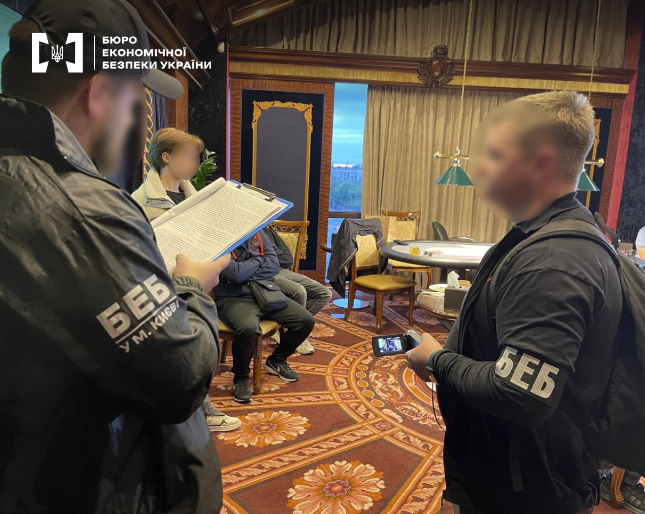 Група людей у розкішному інтер’єрі з класичними меблями та візерунчастим килимом. Детектив БЕБ оголошує підозру. Інший детектив фіксує процес на відео