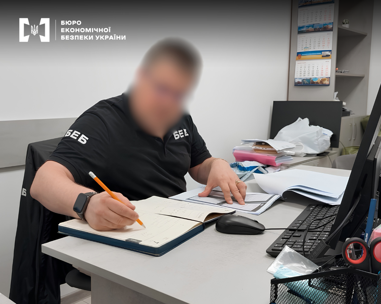 An ESBU detective is taking notes in a logbook at an office desk. The face is blurred, with documents and stationery scattered on the table