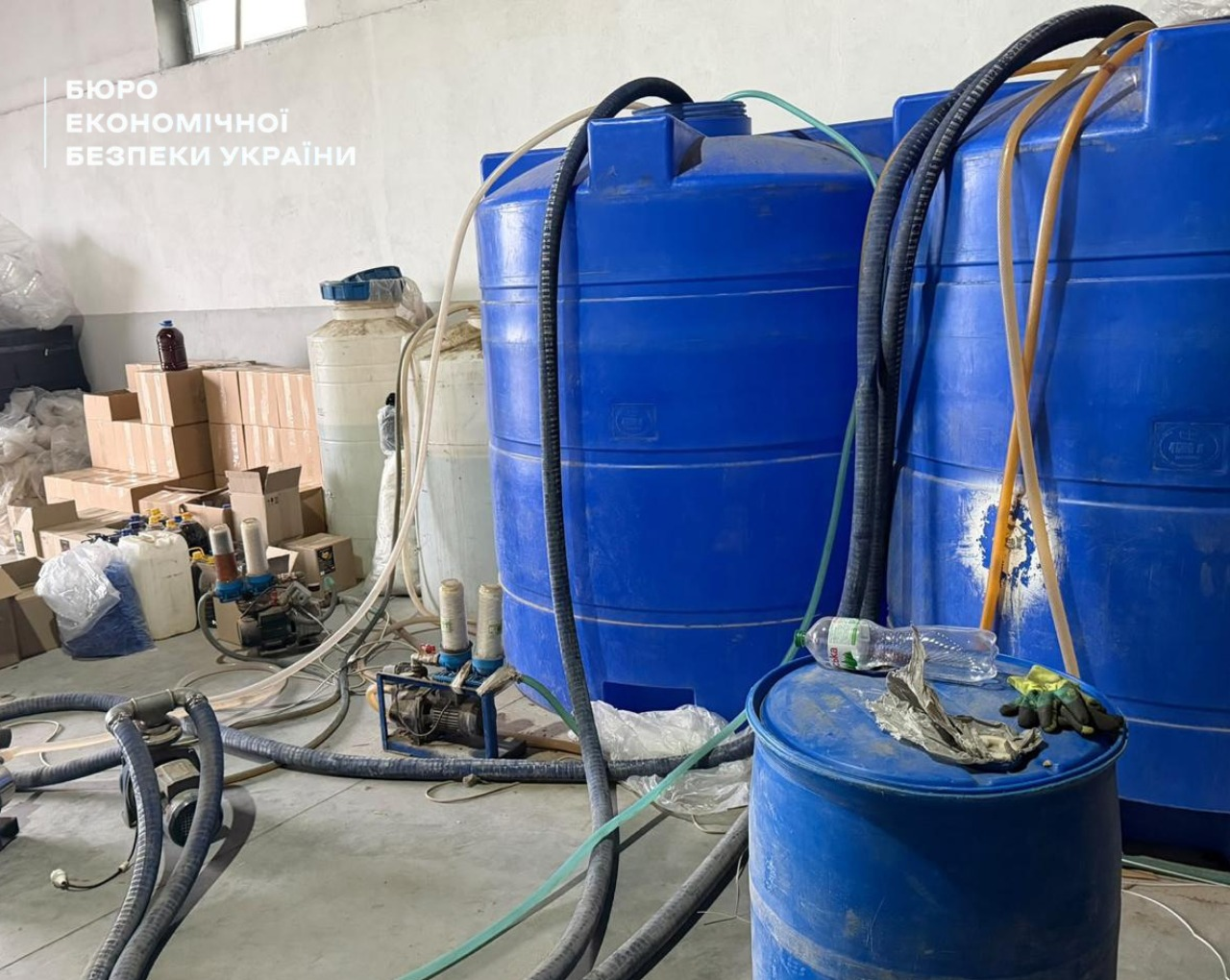 Large blue plastic containers, pumping equipment, connected hoses, and stacks of boxes with finished goods in a large workshop