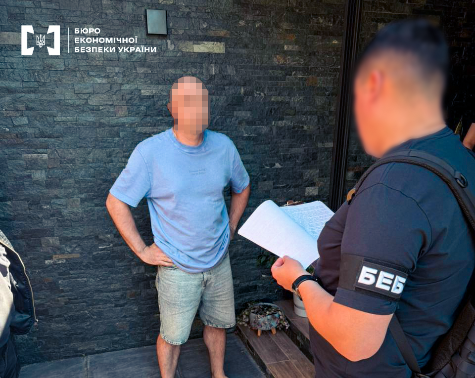 An ESBU detective is reading out procedural documents to a suspect wearing shorts near the entrance to a building. The faces are blurred.