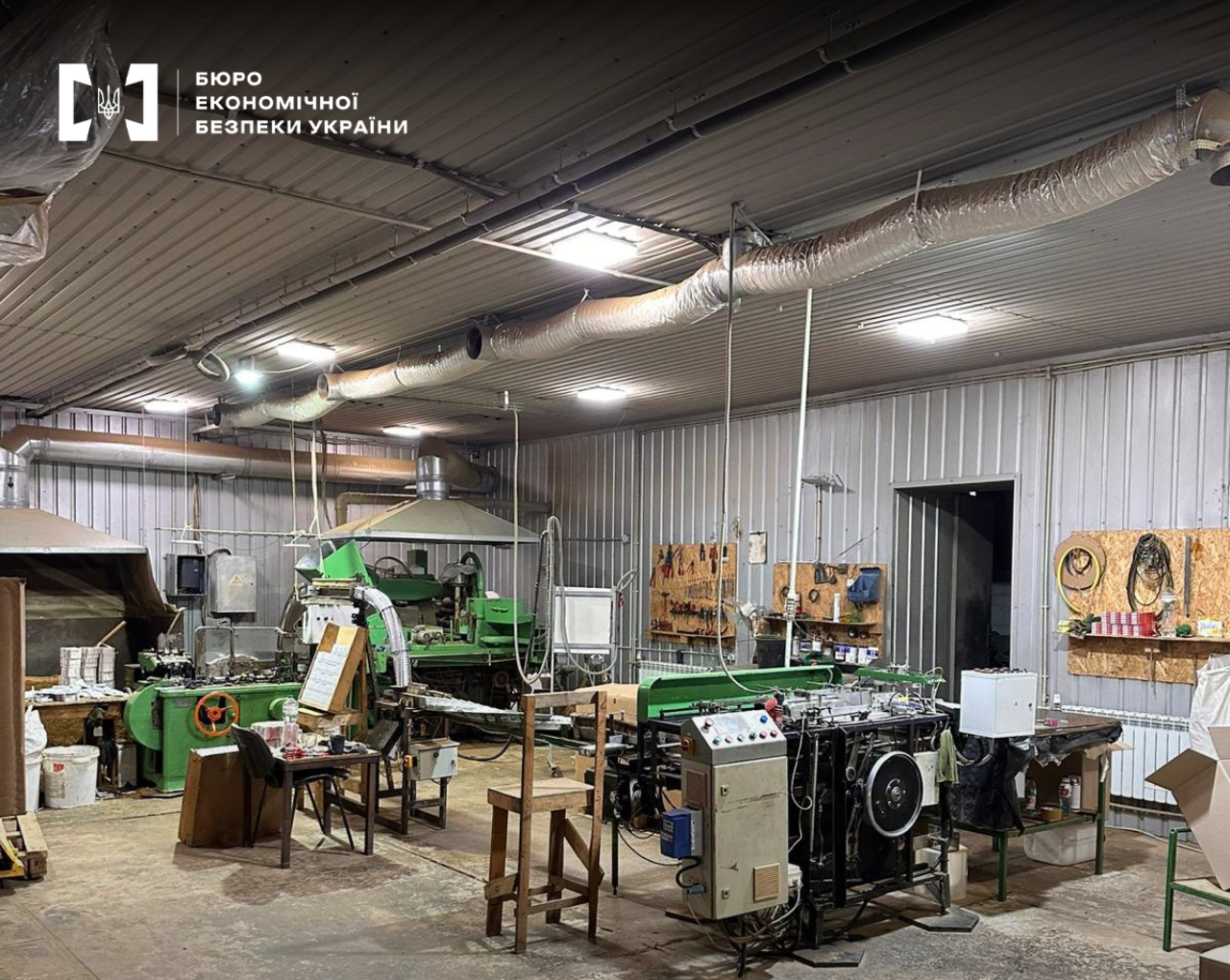 A warehouse converted into a production facility: tobacco manufacturing machines, paper rolls, tools on the walls, and ventilation pipes under the ceiling