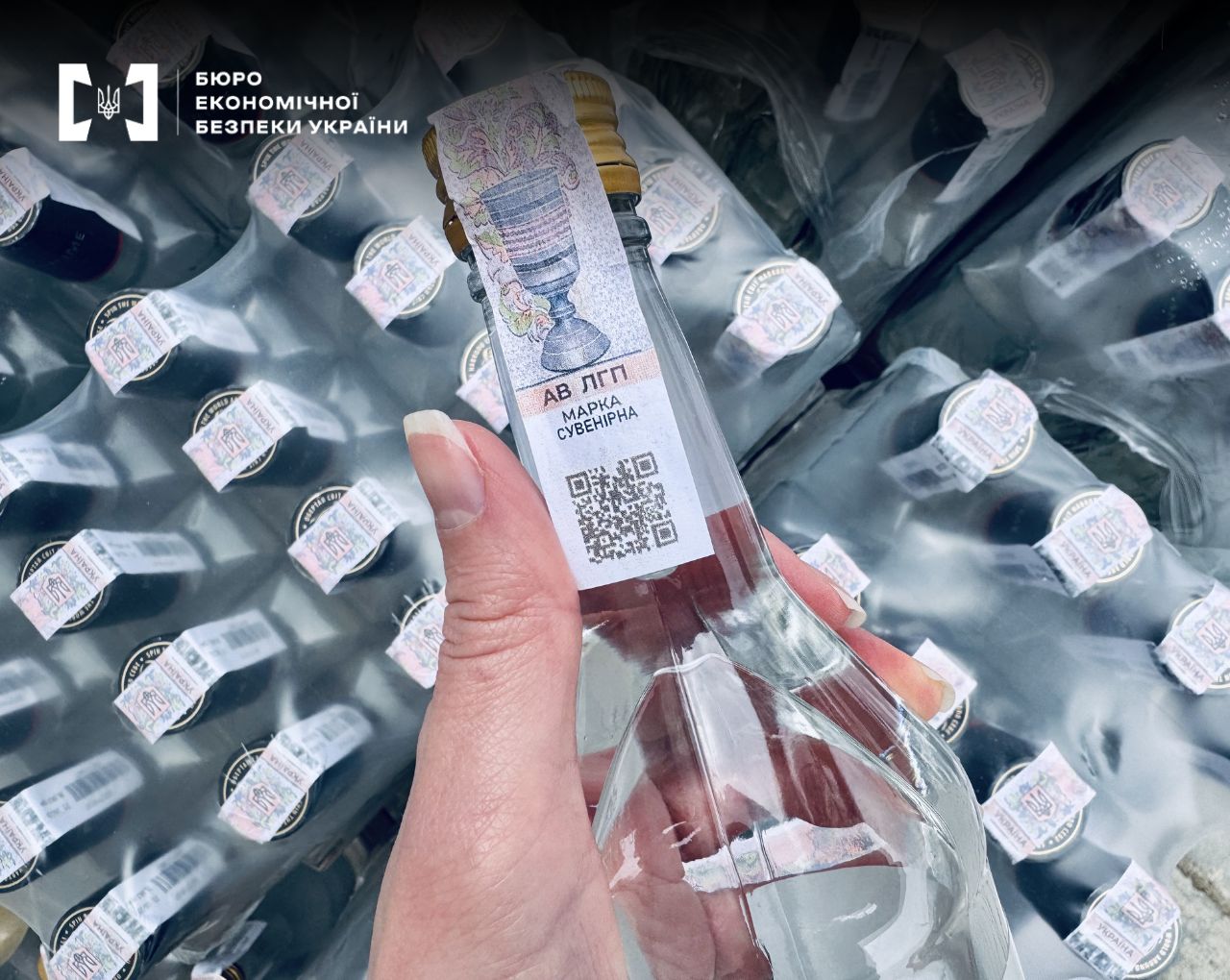 A hand holds a glass bottle containing a clear liquid, with a stamp and a QR code affixed to the neck. In the background, a large number of similar bottles in packaging are visible