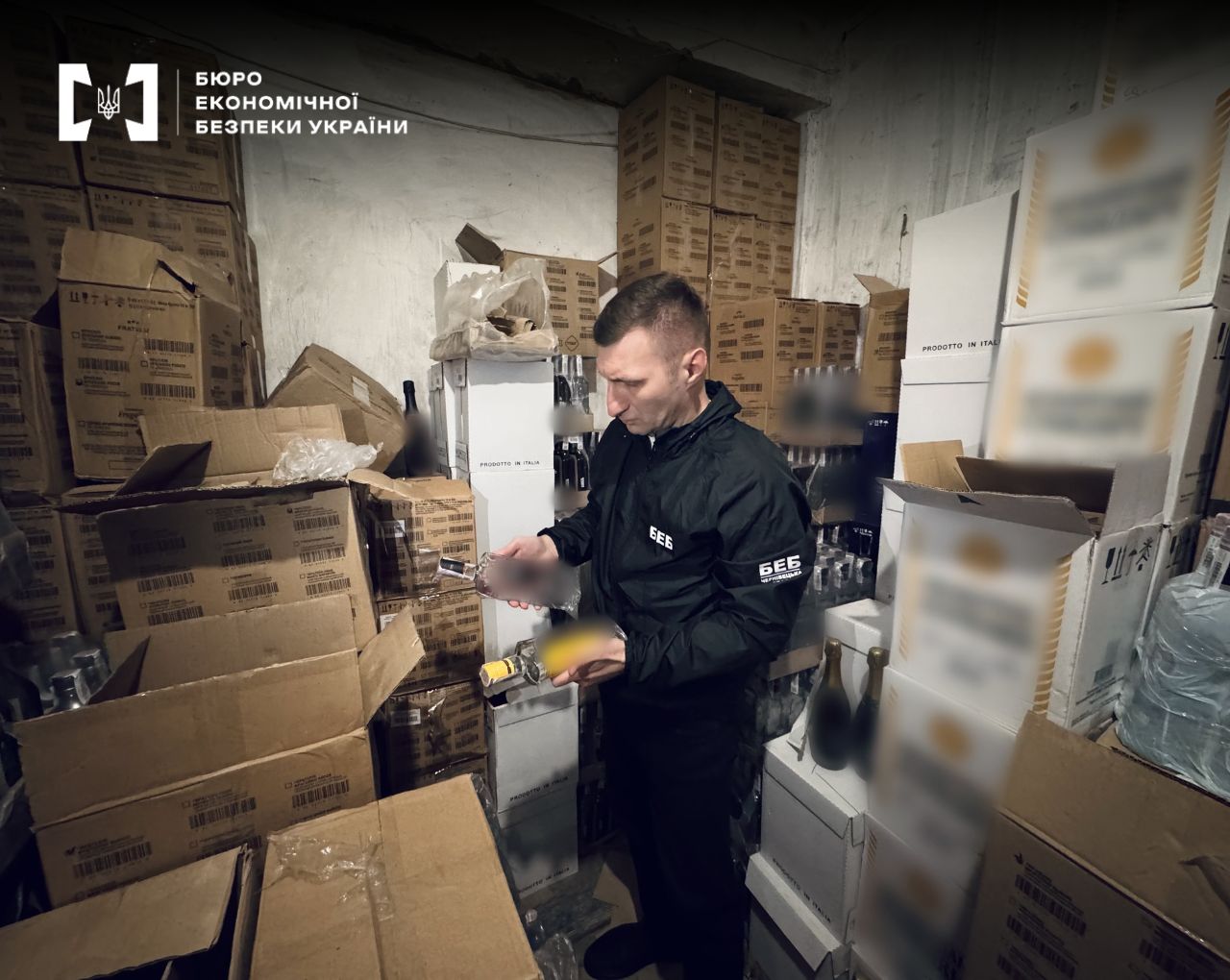 An ESBU detective is holding two bottles of alcohol and examining them. He is surrounded by a large number of cardboard boxes and cases of drinks in a warehouse facility