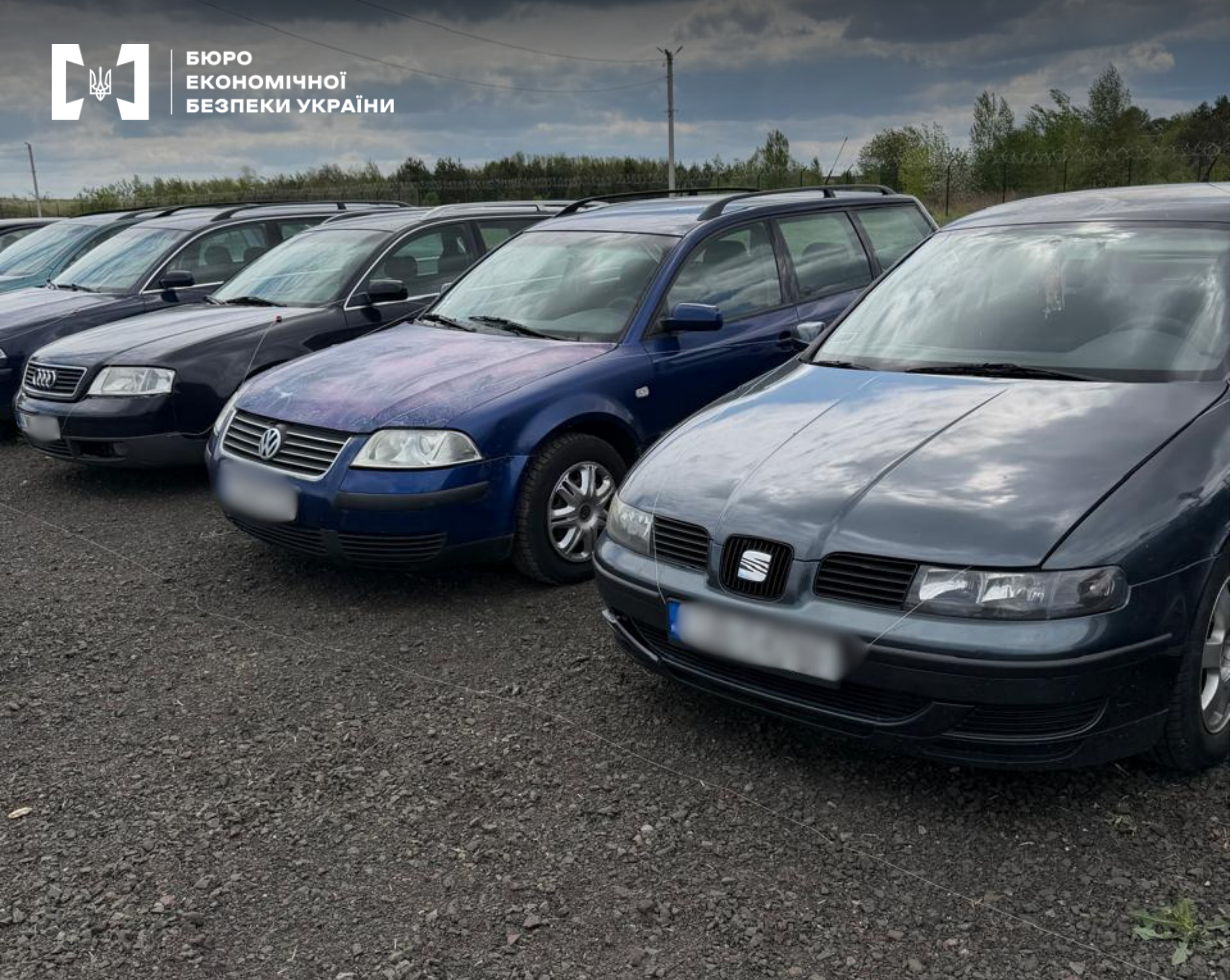 Dark-colored station wagons and sedans are lined up in a single, tight row on a gravel surface outdoors