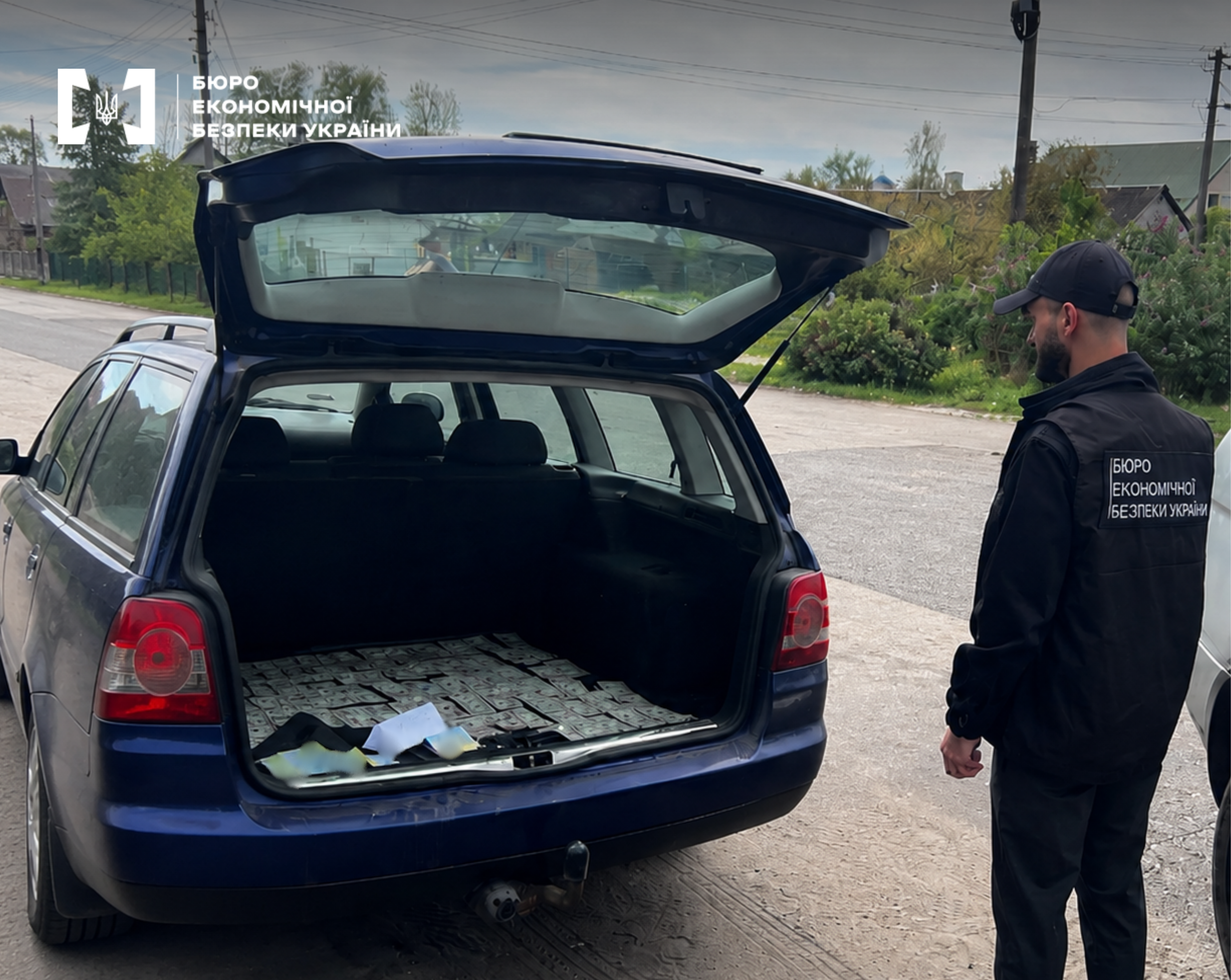A rear view of the open trunk of a dark blue station wagon. Its floor is completely lined with neat rows of US dollars. An ESBU detective stands nearby.