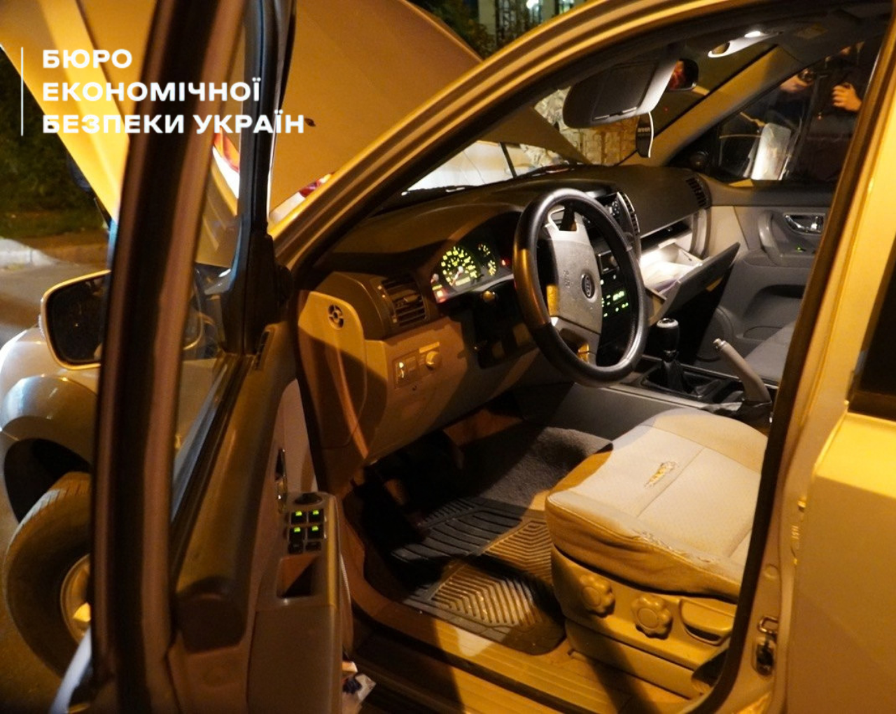 Світло-сірий автомобіль із відчиненими передніми дверима, увімкненою панеллю приладів та підсвіткою салону під час нічного обшуку