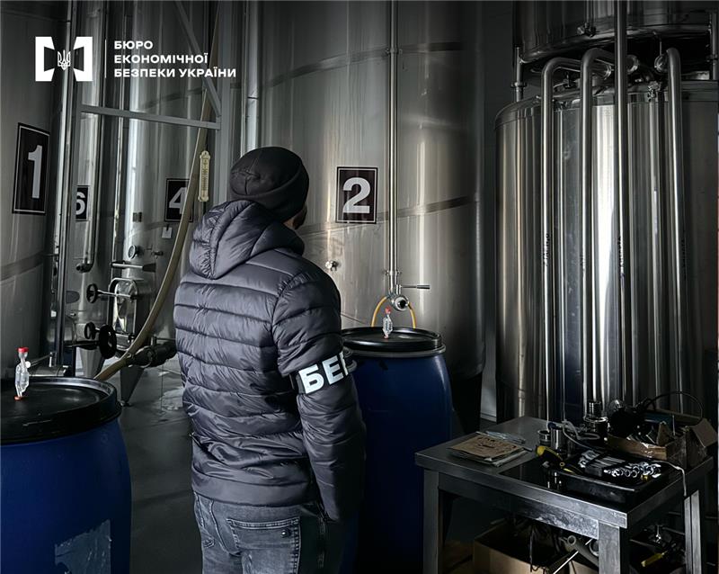 Детектив БЕБ оглядає цех з виробництва напоїв з пронумерованими вертикальними циліндричними металевими резервуарами та синіми бочками