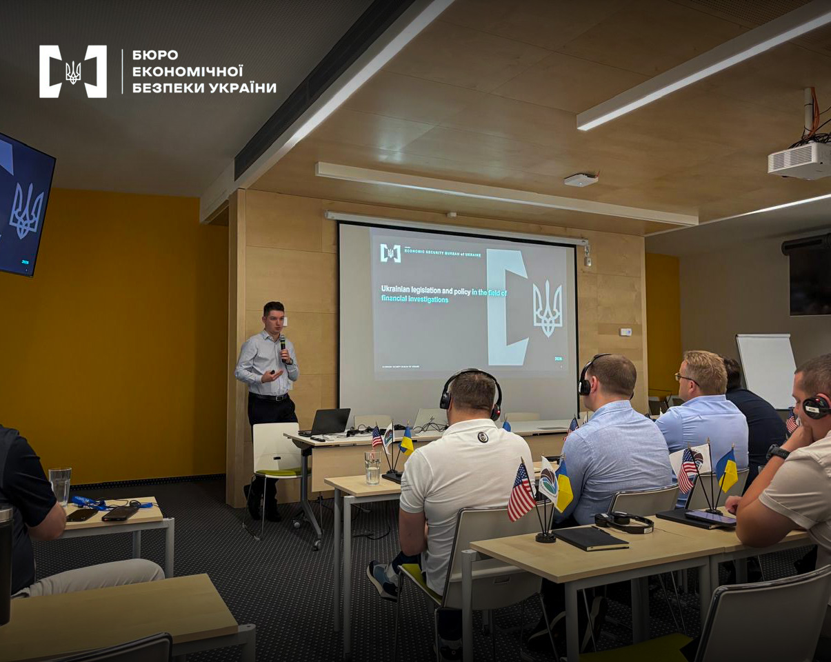 A speaker addresses listeners wearing headphones against the background of the presentation 'Ukrainian legislation and policy in the field of financial investigations'.