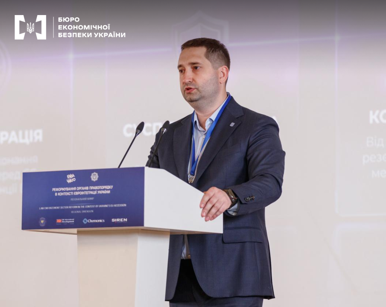 An ESBU representative is delivering a report at a regional forum dedicated to law enforcement sector reform. The speaker stands before microphones against the backdrop of a presentation screen.
