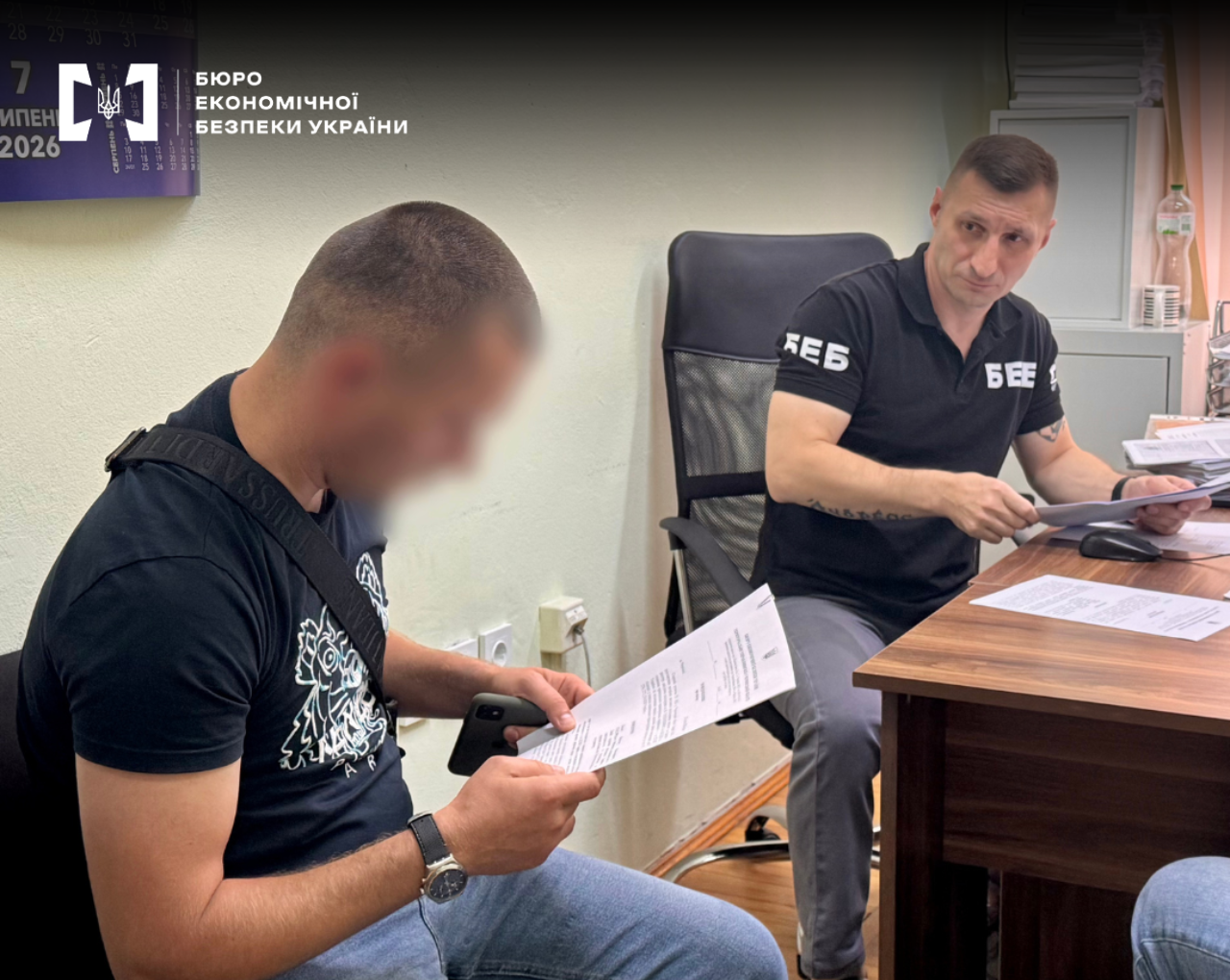 A ESBU detective at a desk provides documents for a man to sign in an office.