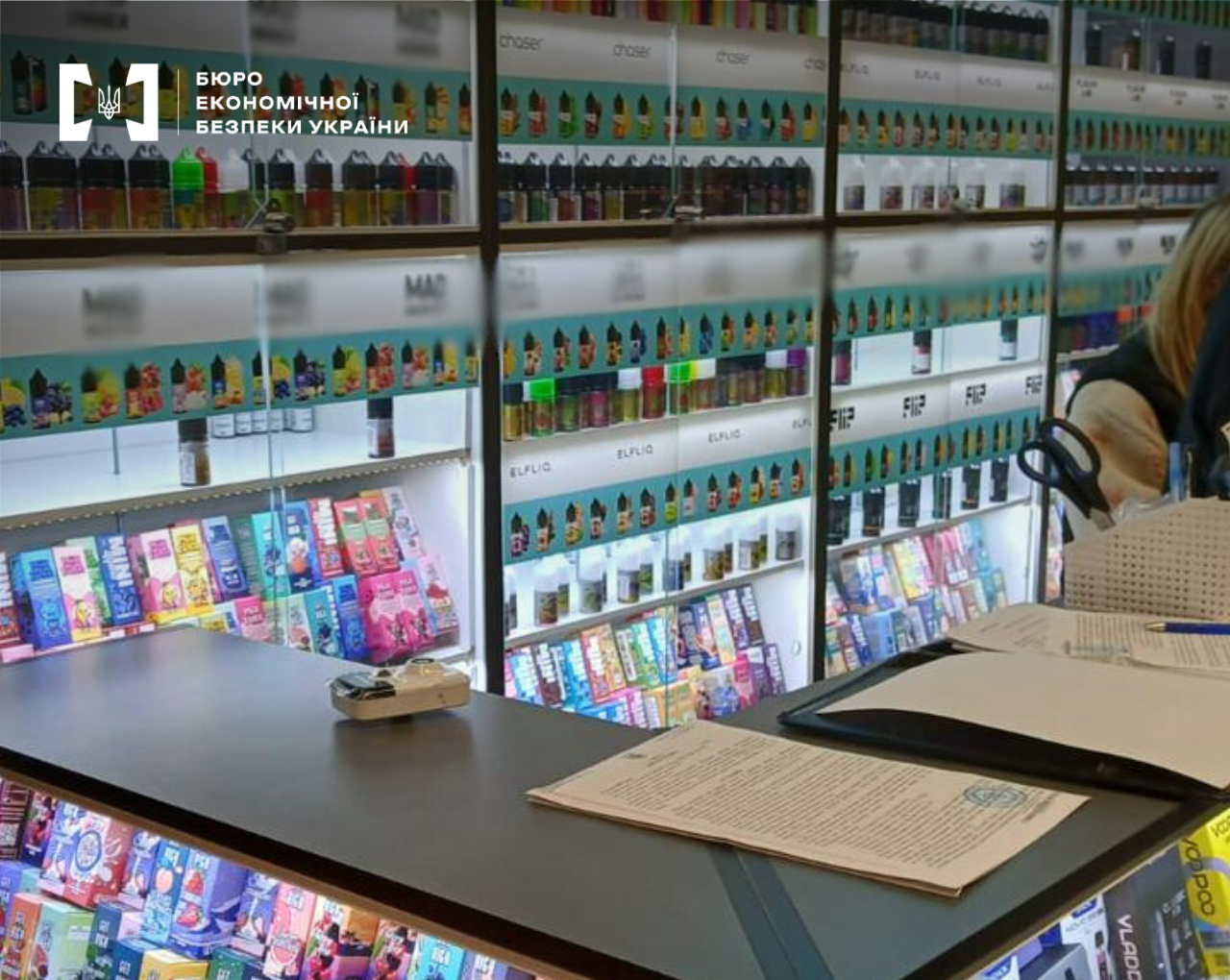 Detectives of the ESBU are conducting a search in an e-cigarette shop. In the foreground is a table with laid-out procedural documents and a blue seal.