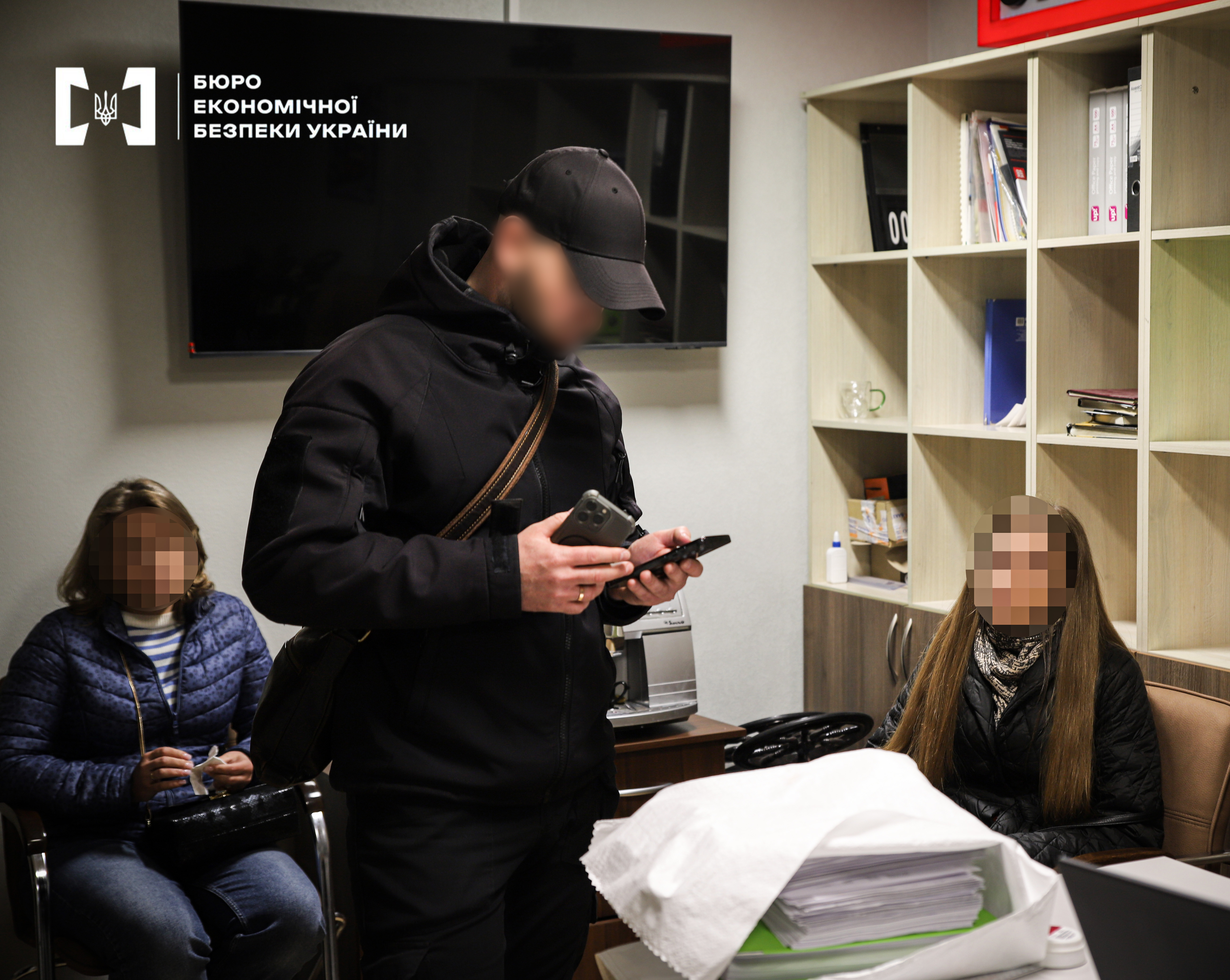An ESBU detective is holding a smartphone while standing in front of two women in an office. A shelving unit with folders and documents stands nearby. The people’s faces are blurred